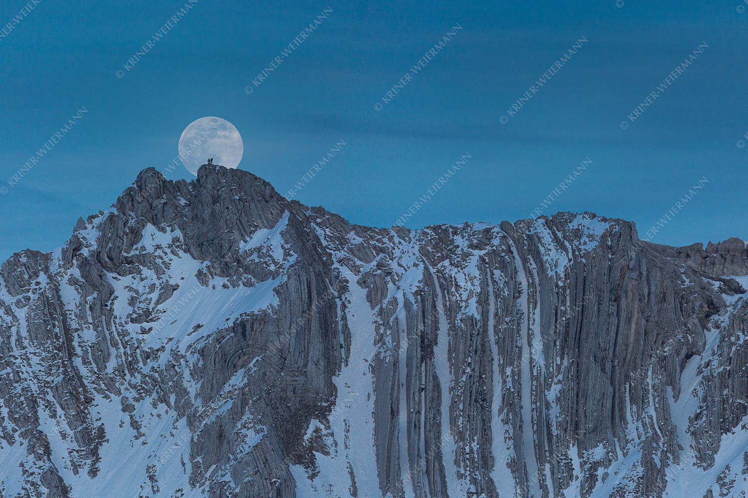 Der Vollmond wirkt riesig im Vergleich zu den beiden Menschen auf dem Gipfel